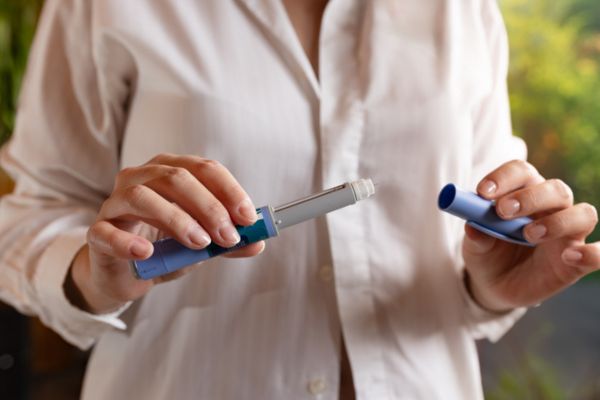 Woman in white shirt, standing in a rustic living room, is holding semaglutide injection pen in her hands.
